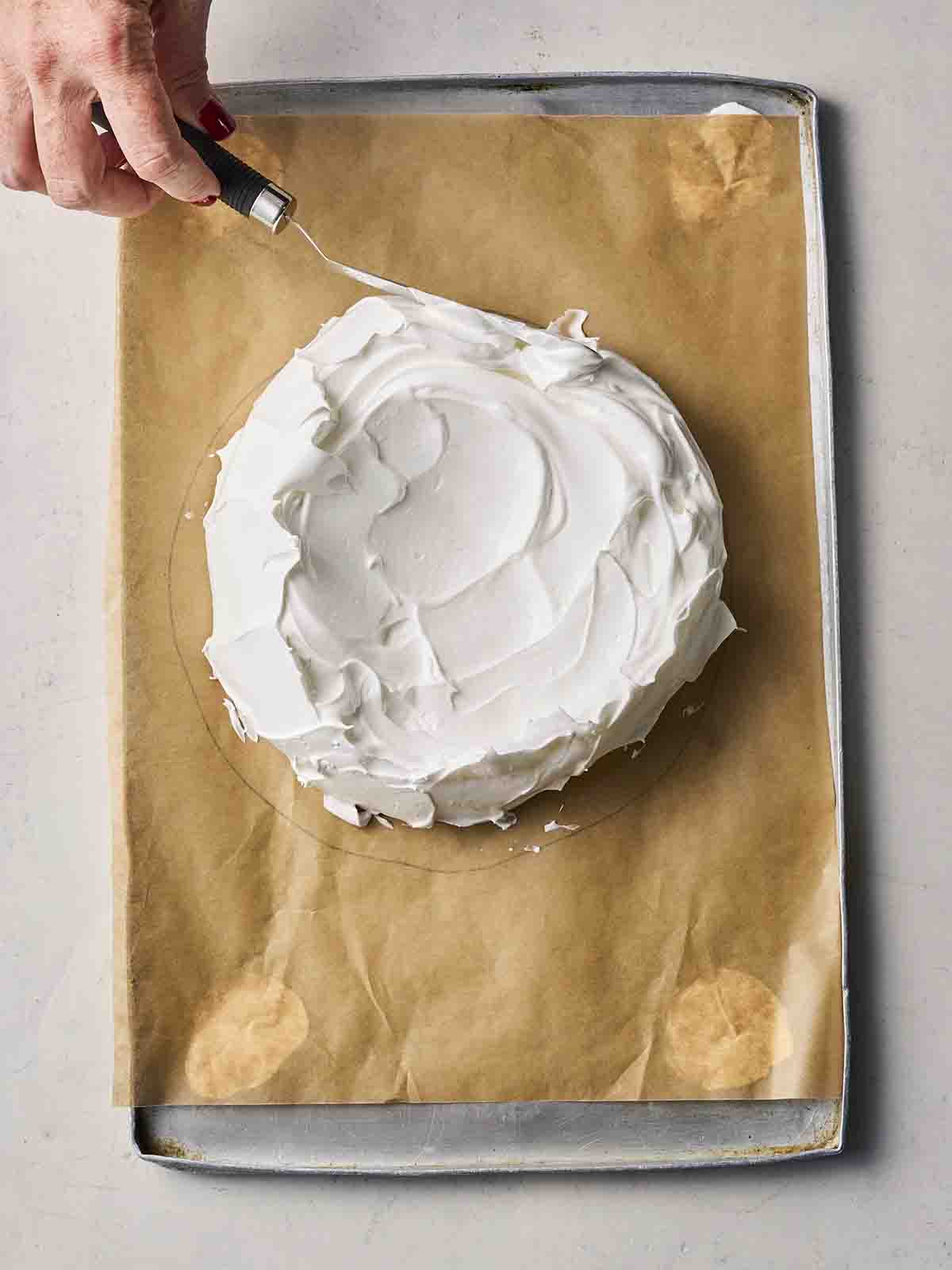 A hand with a palette knife smoothing the edges of a meringue on a baking tray.