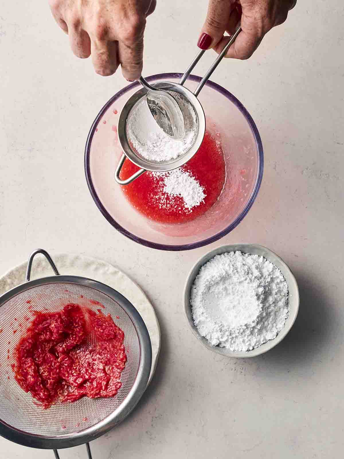 A hand pushing icing sugar through a sieve into a glass bowl of raspberry juice, with other bowls to the side.