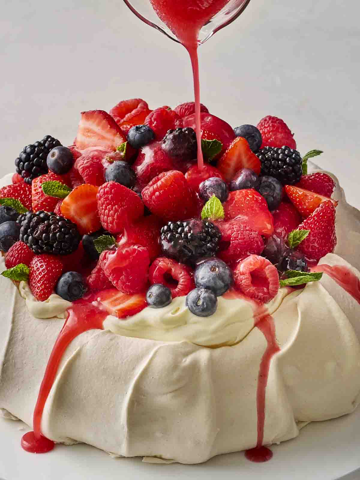 A pavlova with fresh fruit on top, with a jug pouring a red juice over the top.
