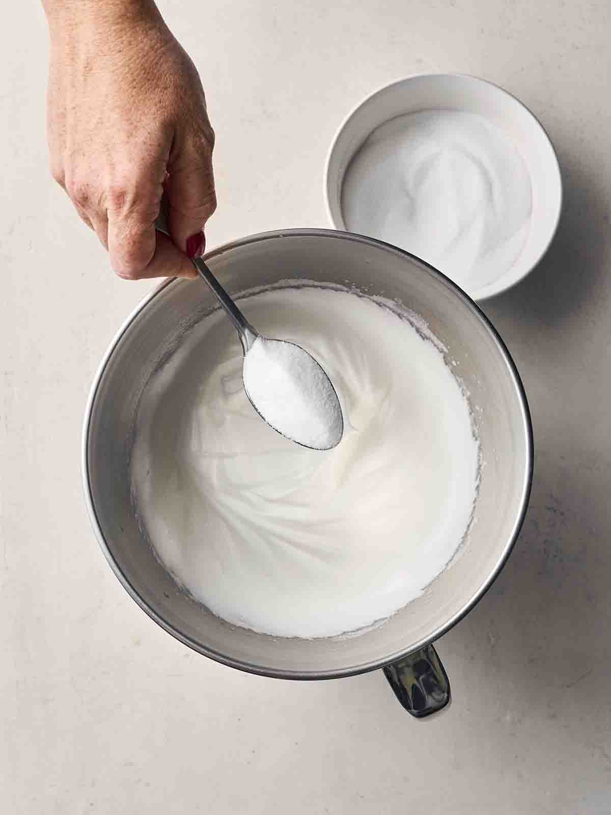 A large mixing jug with egg whites inside with a hand about to add a spoonful of icing sugar.