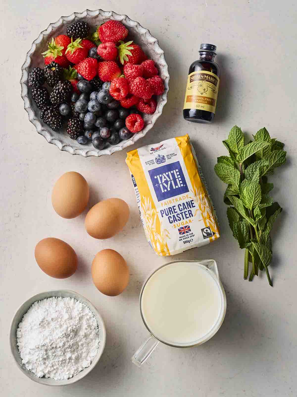 Ingredients laid out on a counter, including fresh berries, four eggs, caster sugar, mint, vanilla, icing sugar and cream.