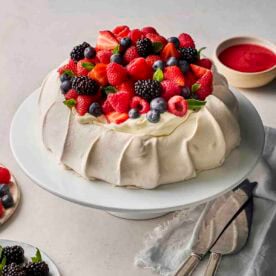A homemade pavlova dessert, on a cake stand, topped with whipped cream and fresh berries, with raspberry sauce in the background.