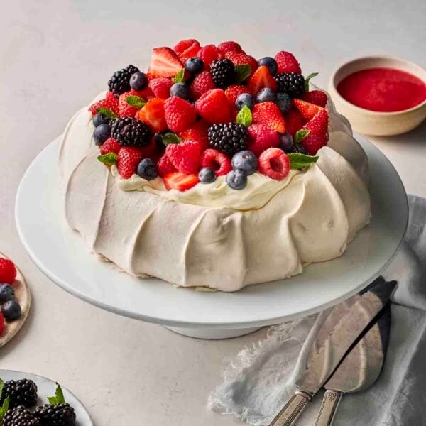 A homemade pavlova dessert, on a cake stand, topped with whipped cream and fresh berries, with raspberry sauce in the background.