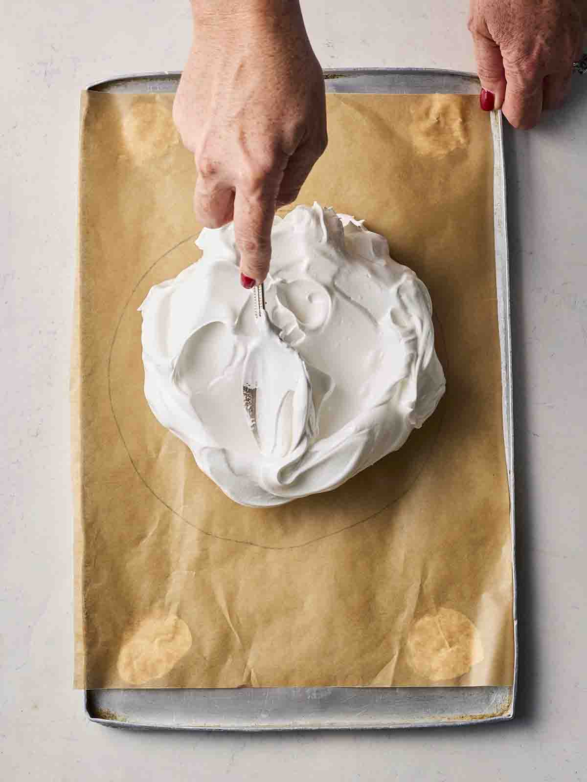A hand spreading meringue mixture onto a baking sheet with baking paper and a drawn-out circle.