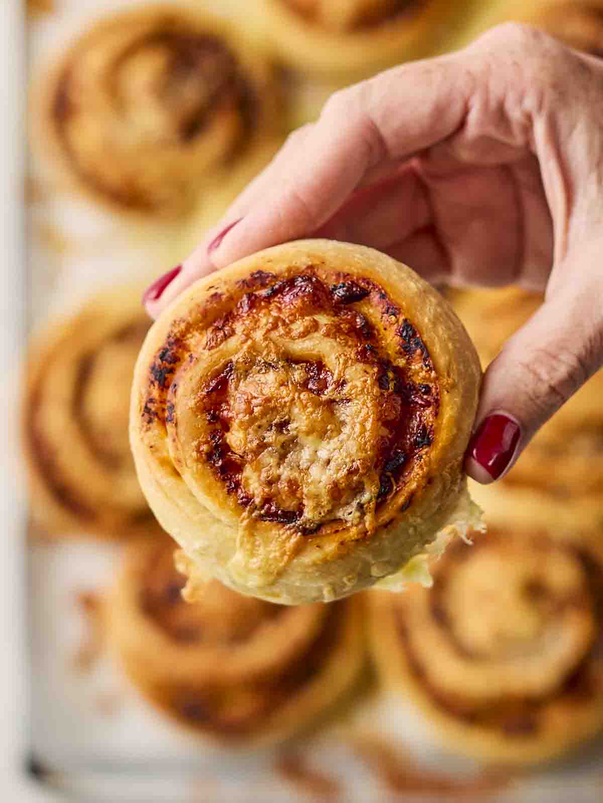 A hand holding a baked pizza roll, with lots more on a tray in the background.