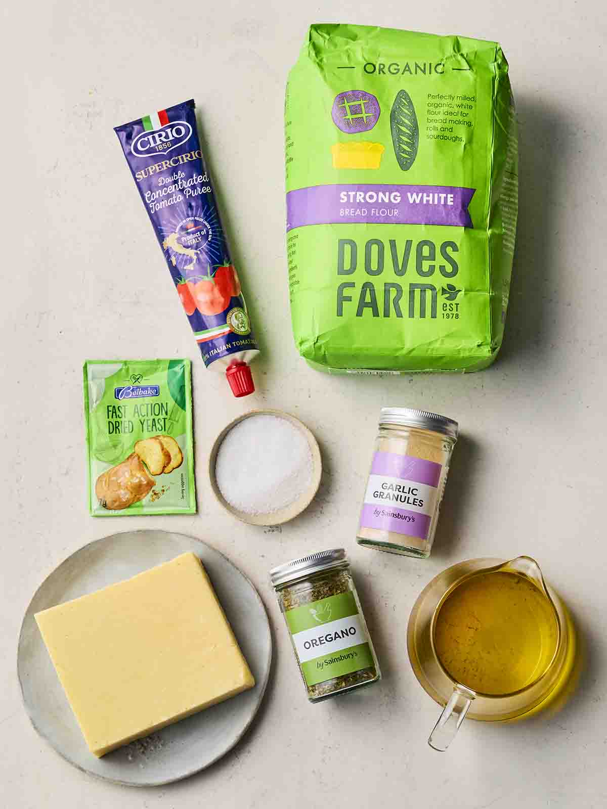 Ingredients laid out on a white counter, including strong white flour, tomato puree, dried yeast sachet, oil, cheese, salt and herbs.