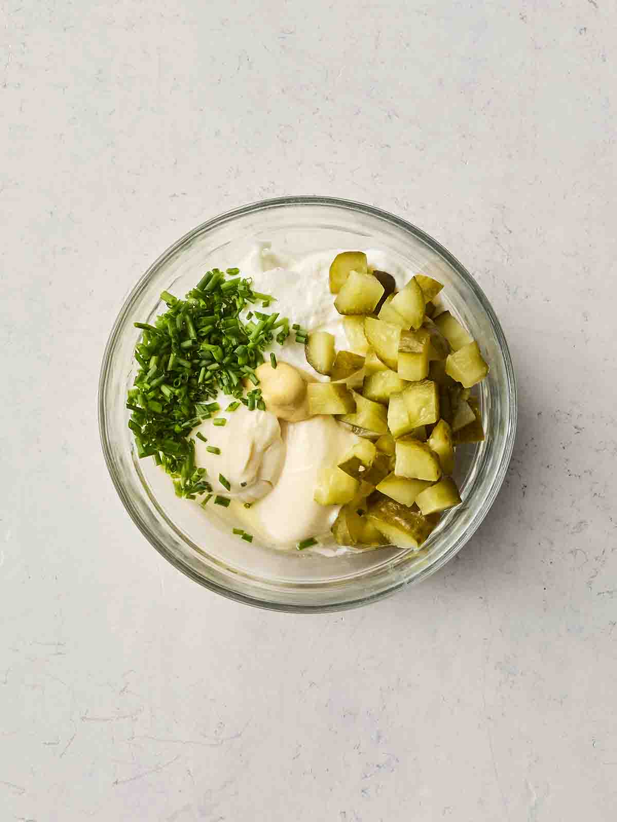 Ingredients ready to be mixed in a glass bowl, including gherkins, mayonnaise, yoghurt, chives and garlic.