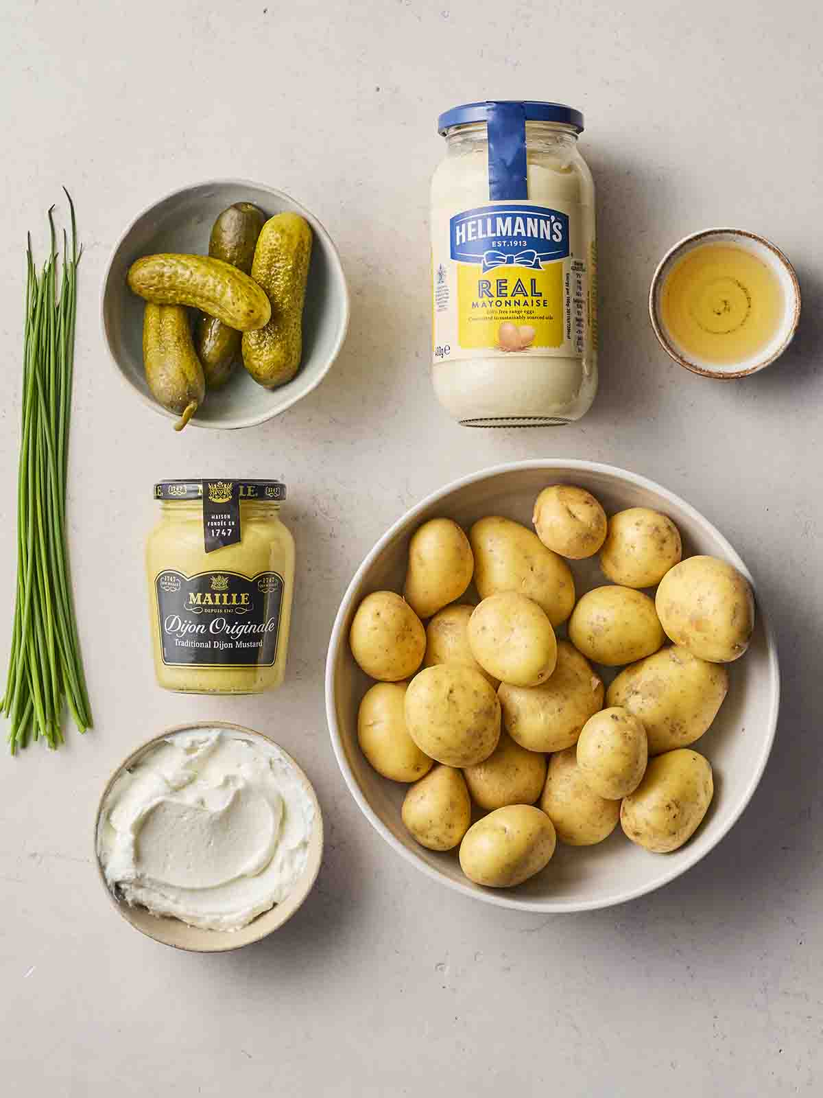 Ingredients laid out on a white counter, including potatoes, gherkins, mayonnaise, chives, mustard and yoghurt.