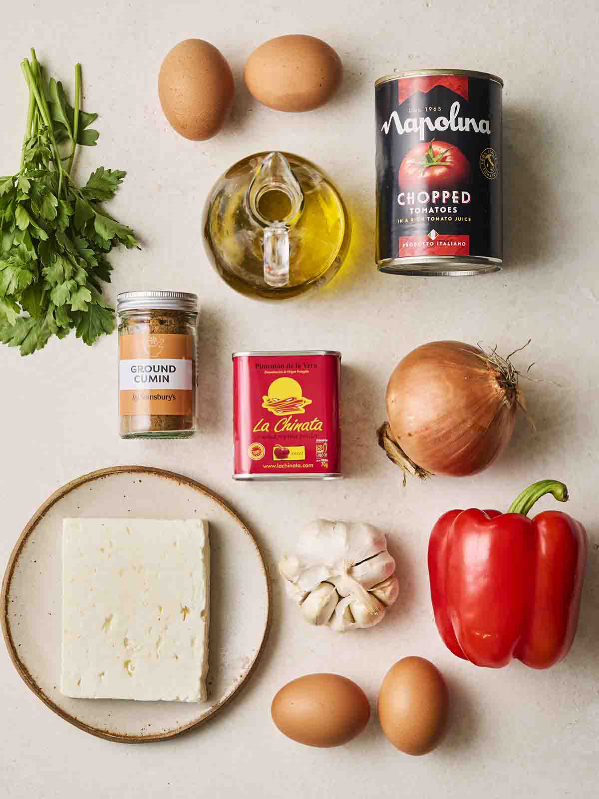 Ingredients on a white counter, including tinned tomatoes, eggs, onion, oil, feta, pepper and garlic.