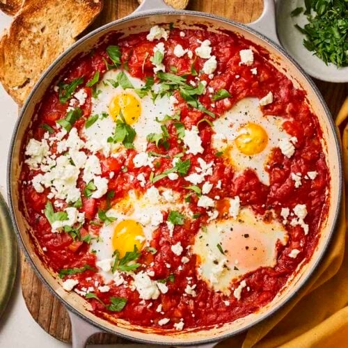A pan on a wooden board, with eggs and tomatoes, with toast to the side and a bowl of parsley.