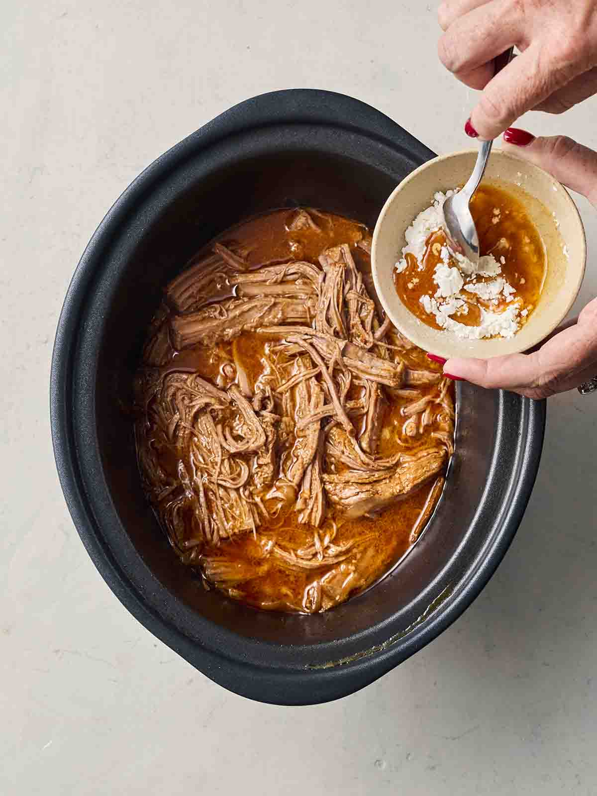 A slow cooker pan with shredded beef in with a sauce, with a hand stirring sauce and cornflour in a small pot over it.