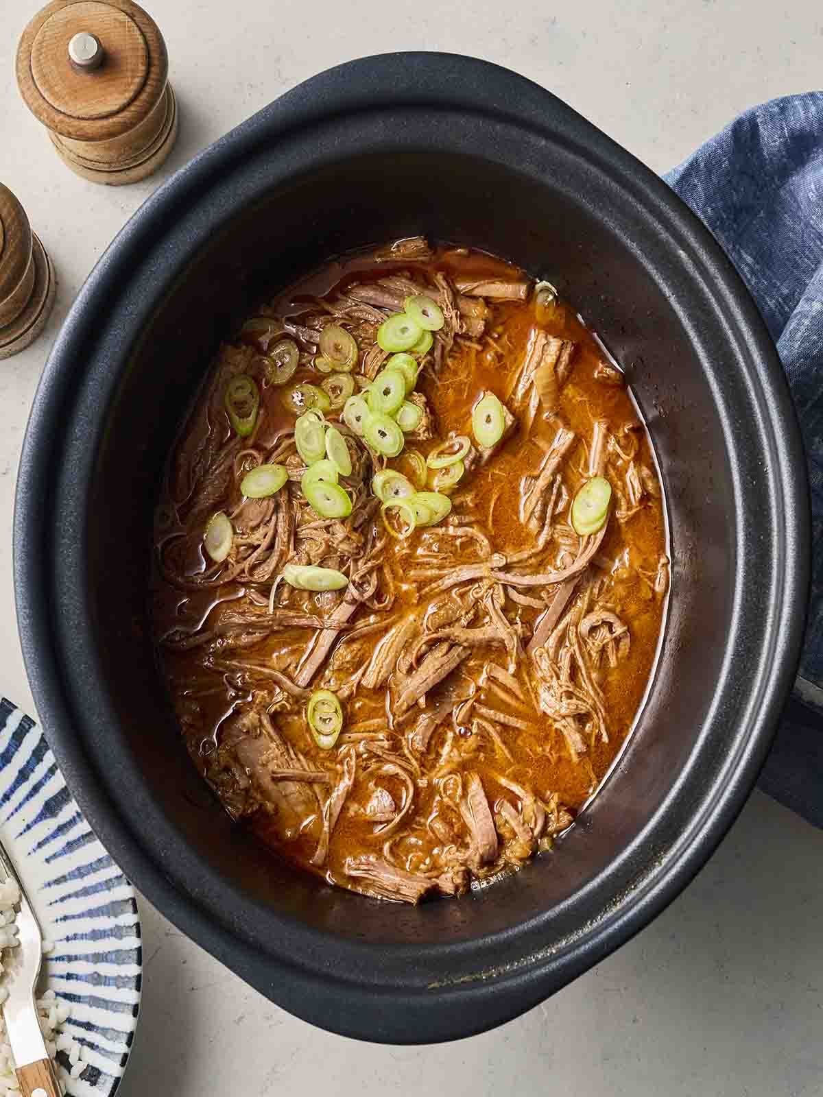 A slow cooker pan filled with cooked Korean Beef Brisket, shredded and topped with spring onions.