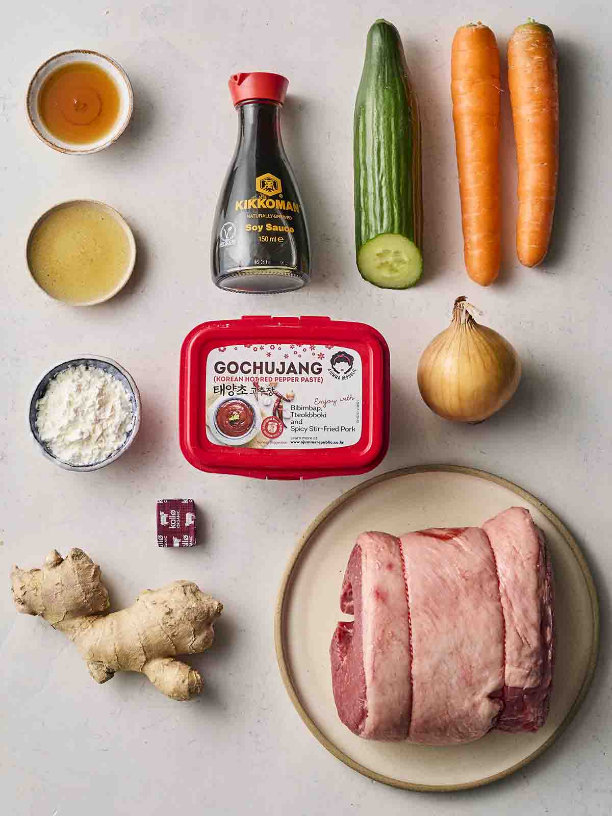 A collection of raw ingredients on a white counter top, including a beef brisket joint, carrots, cucumber, onion, ginger and various Korean-style sauces.