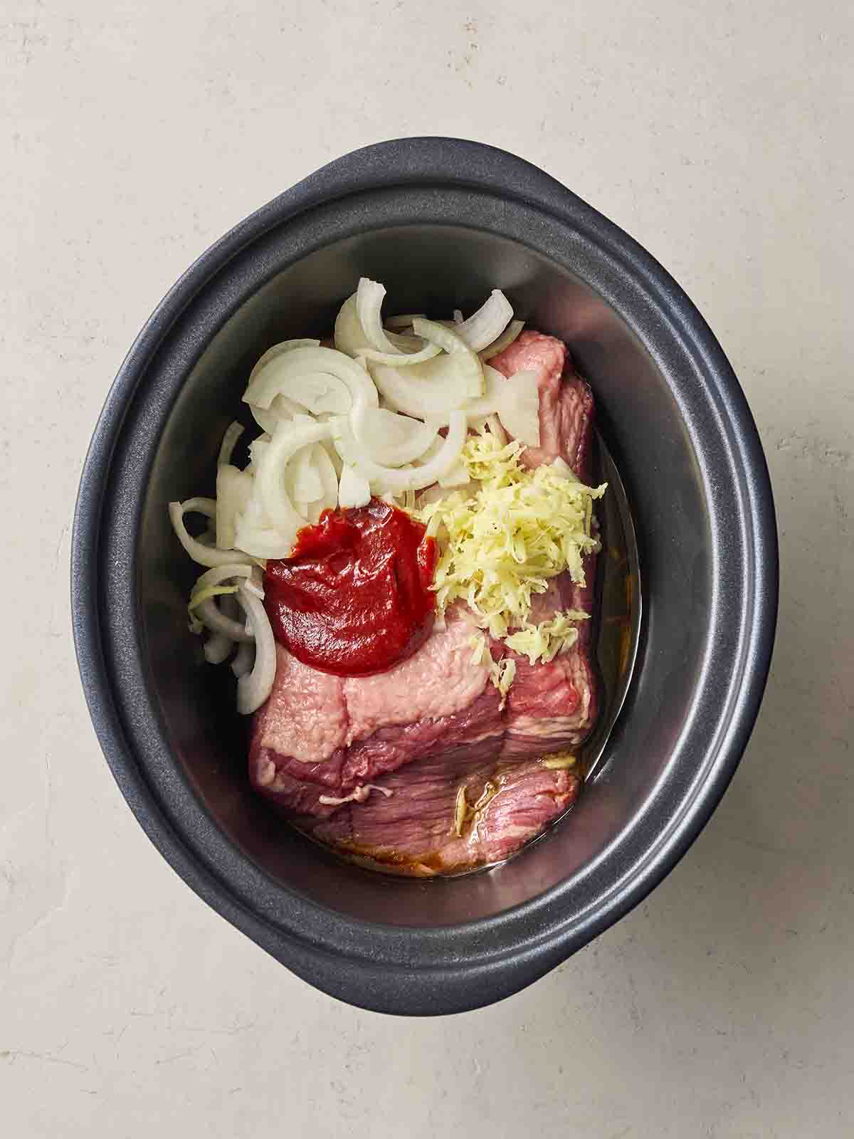 Raw beef brisket with other ingredients, including onion, sauce and ginger, in a black slow cooker pan.