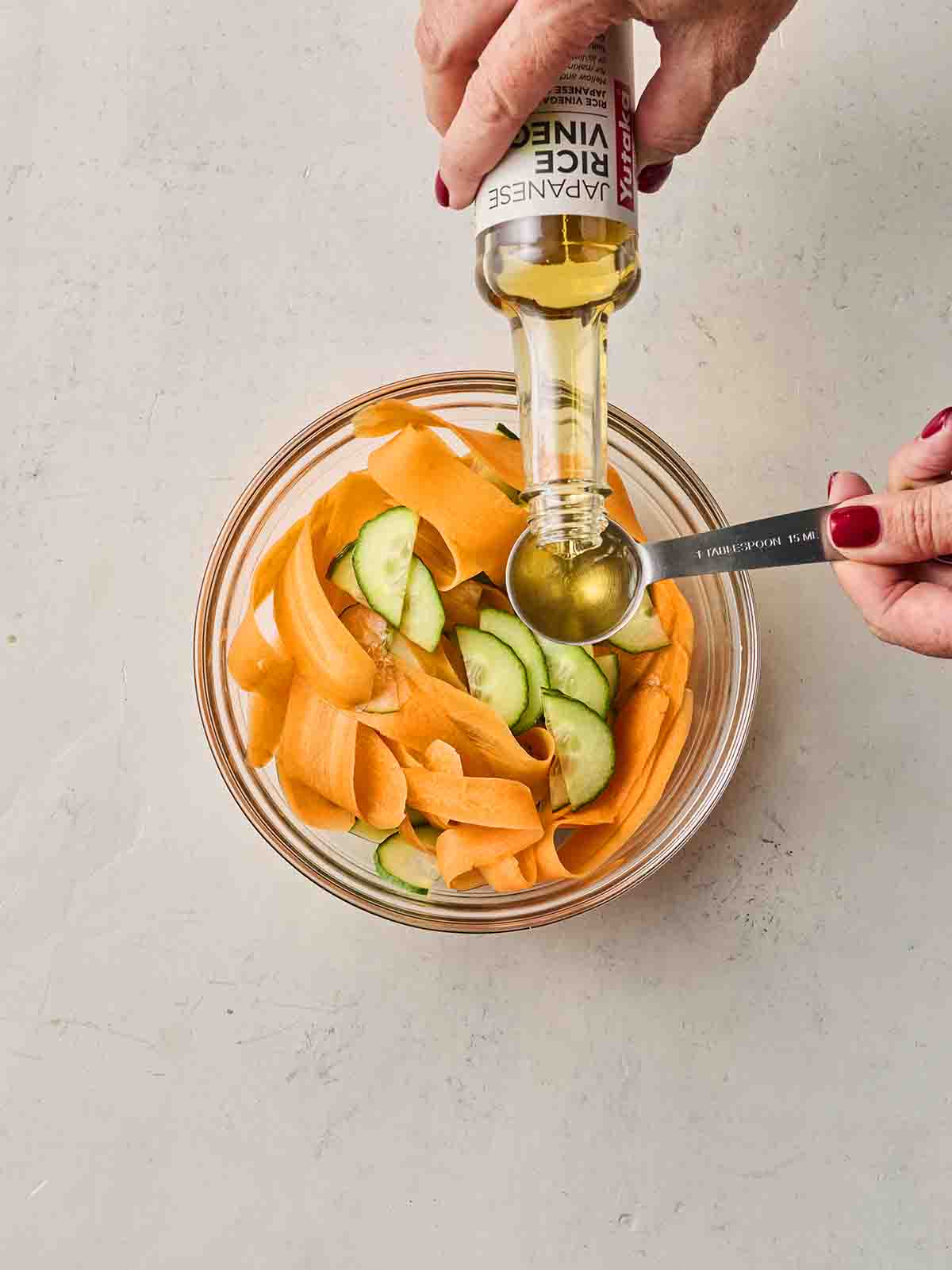 A small glass bowl with shredded carrots and chopped cucumber, with hands pouring rice vinegar into a measuring spoon above it.
