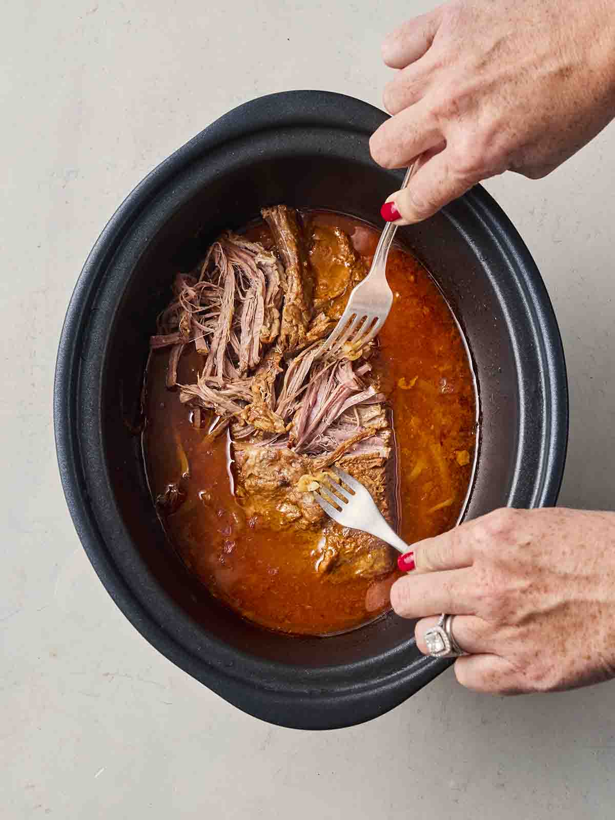 Two hands with two forks, shredding beef in a sauce inside a slow cooker pan.
