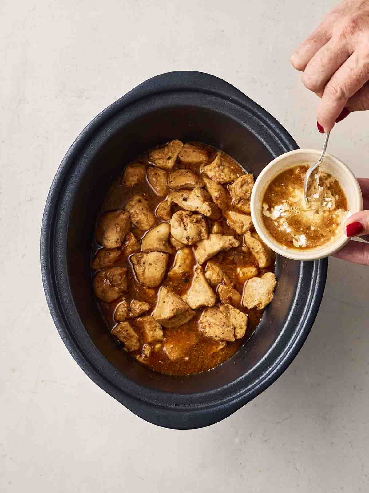 A chicken dish in sauce in a slow cooker, with two hands mixing cornflour and sauce into a small bowl above it.