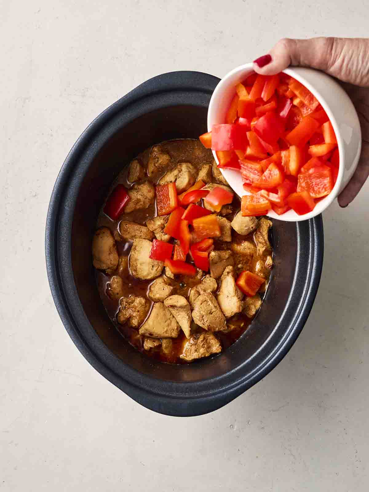 Chunks of red pepper being tipped into a slow cooker chicken dish from a bowl.