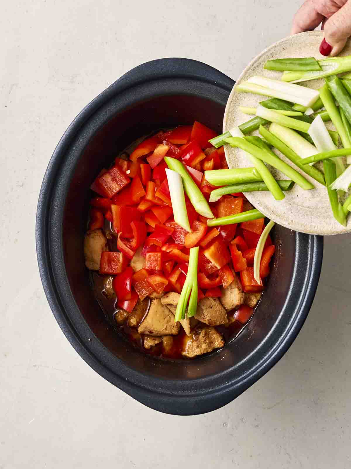 Spring onions being tipped into a slow cooker pan filled with a chicken dish.