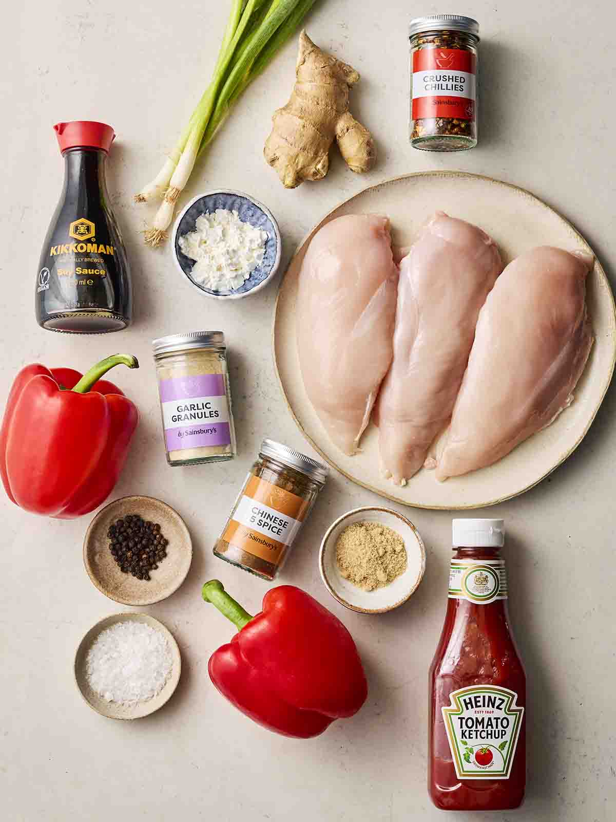 Raw ingredients on a counter top, including chicken breasts, red pepper, ketchup, ginger, soy sauce and other dried spices.