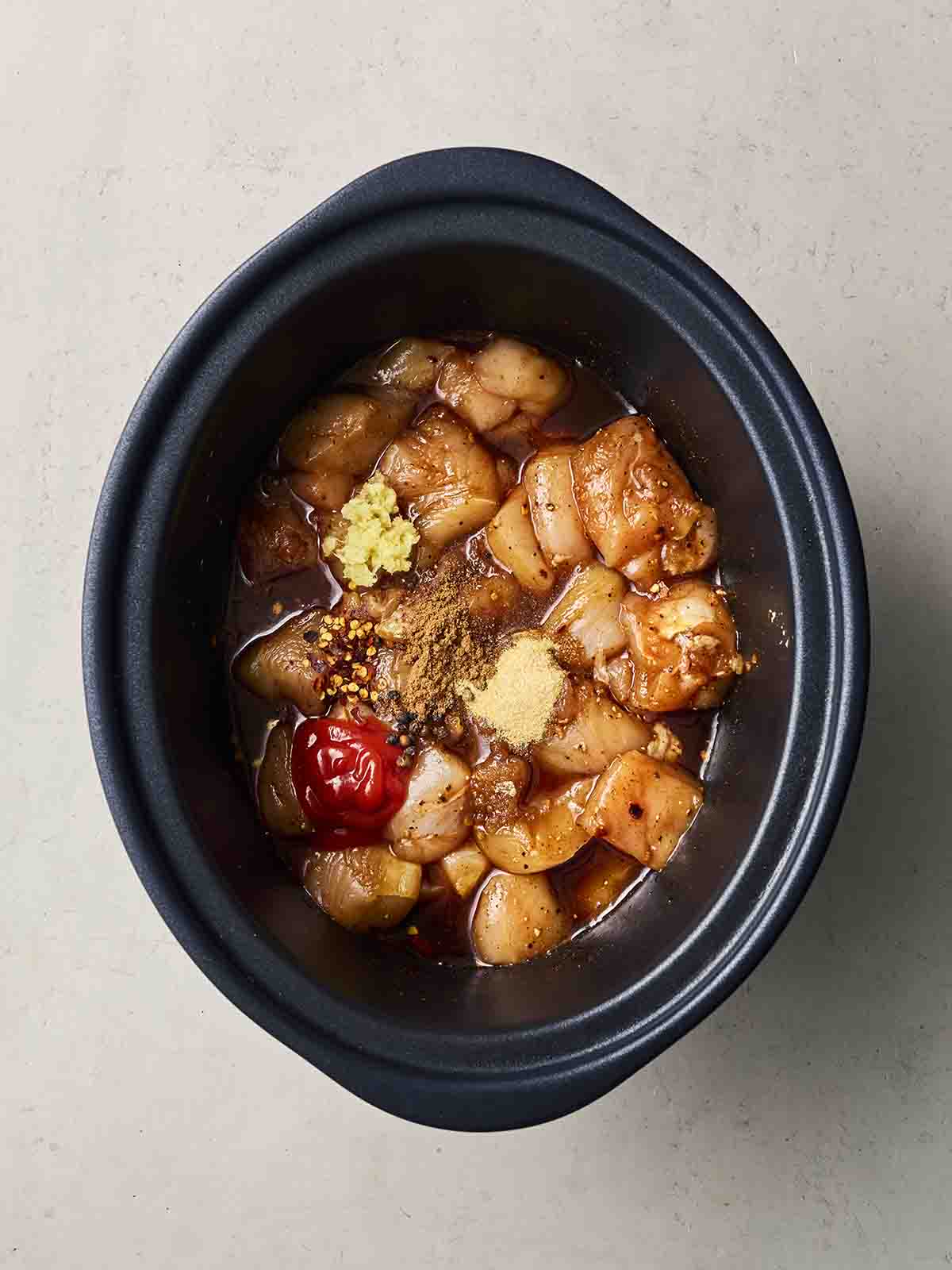 A black slow cooker with raw ingredients inside, including diced chicken, ketchup and spices.