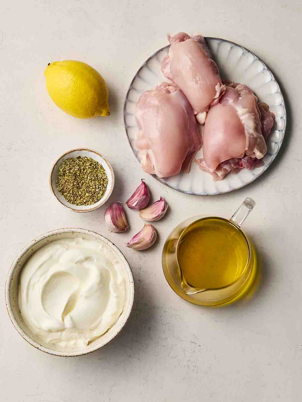 Ingredients laid out on a white counter, including raw chicken thighs, a lemon, oil in a jug, a bowl of yoghurt, garlic and oregano.