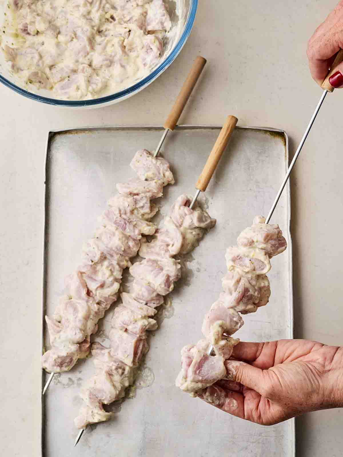Chicken thigh pieces in a yoghurt marinade being threaded onto metal skewers with two hands.