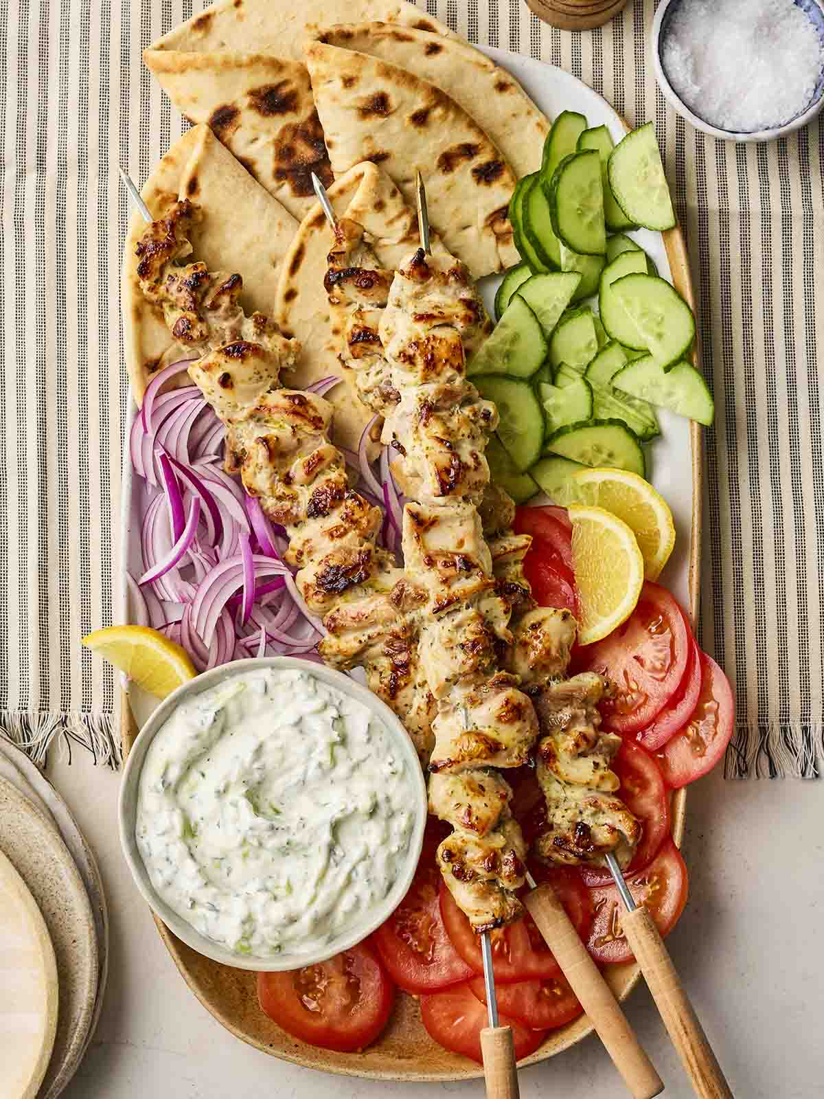A platter with flatbreads, cucumber, chicken souvlaki skewers, red onion, tomatoes and a bowl of tzatziki laid out on a table.