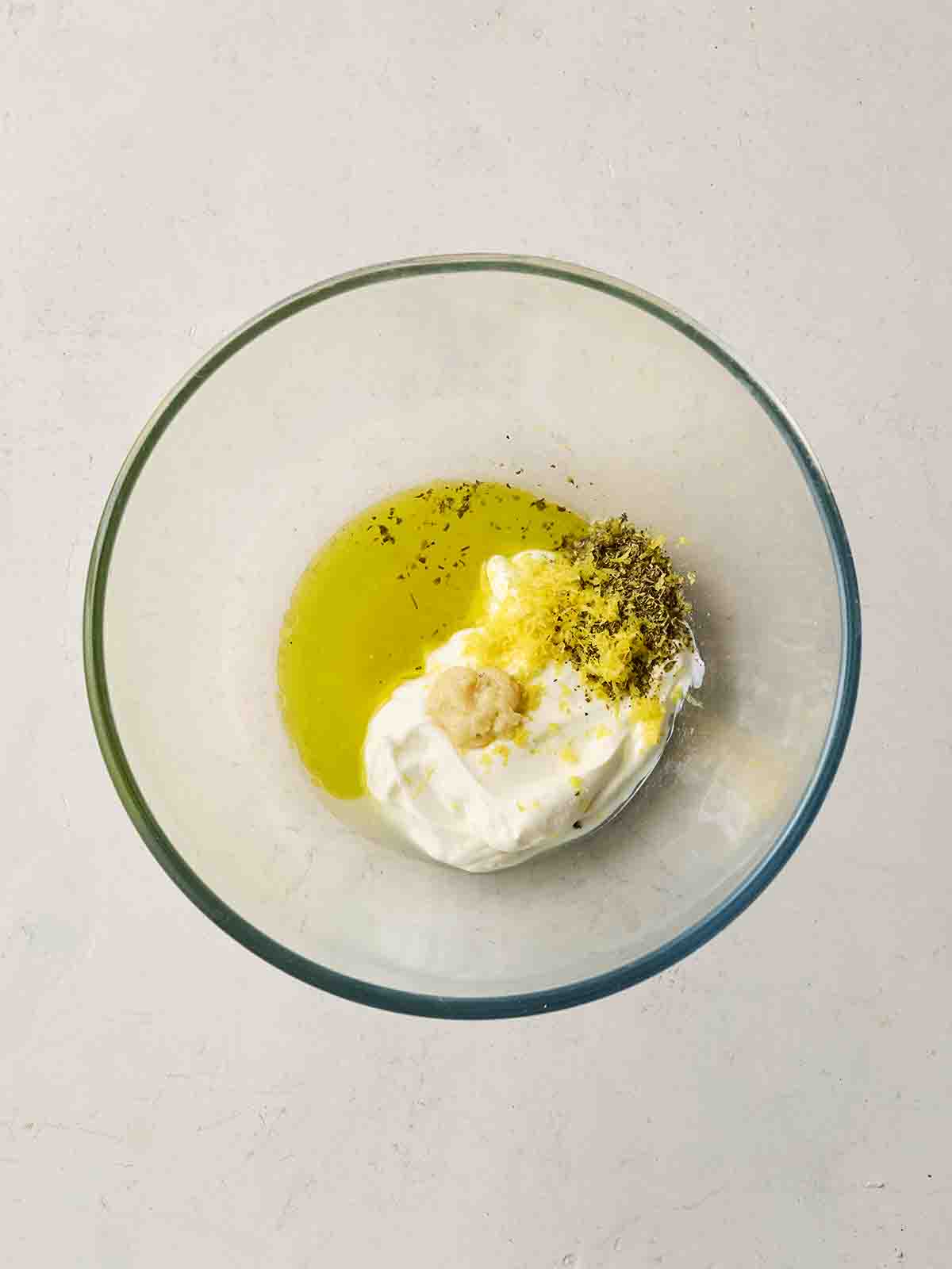 A glass bowl with unmixed olive oil, yoghurt, garlic, lemon zest and oregano in.