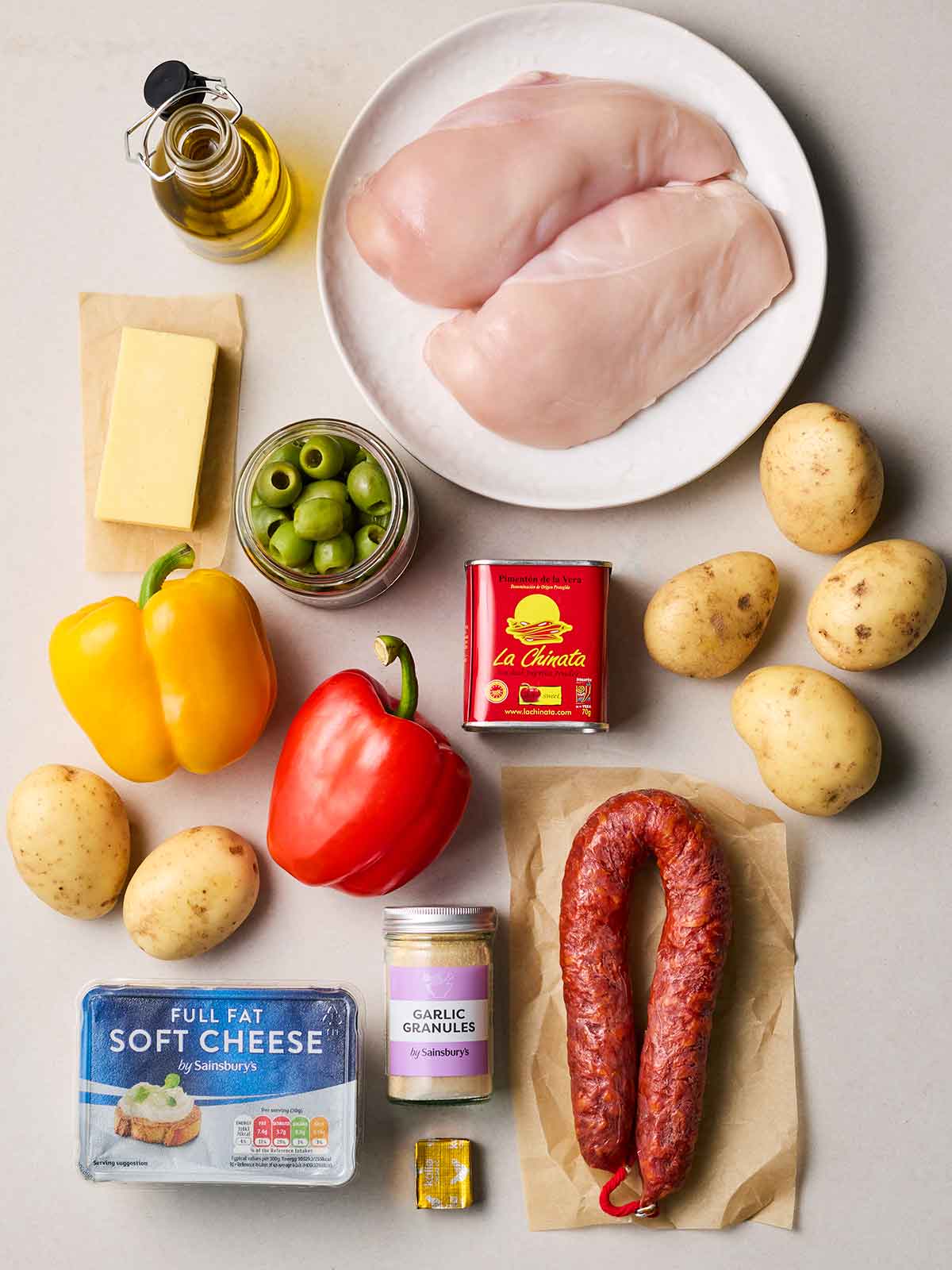 Raw ingredients laid out on a white counter, including chicken breasts, potatoes, chorizo, peppers, soft cheese and others.