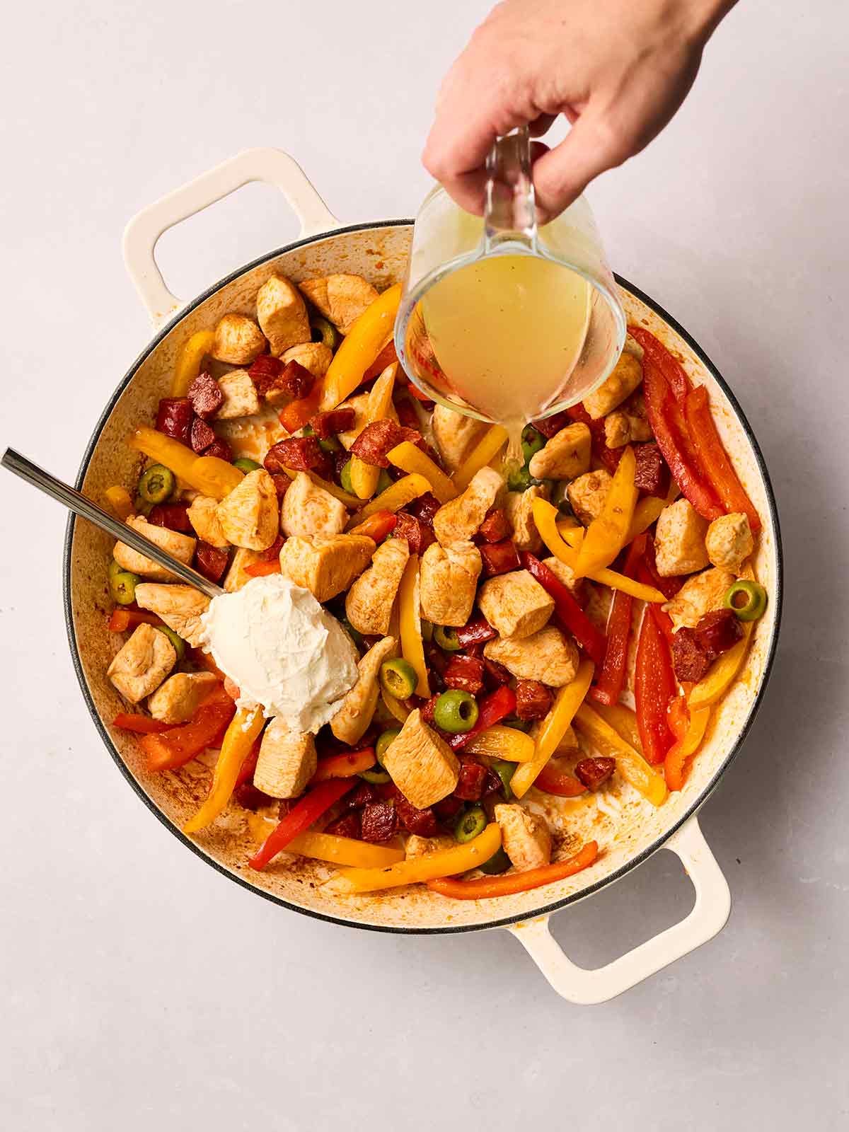 A pan of chicken and chorizo and veg with a spoon stirring in cream cheese and a hand pouring in stock.