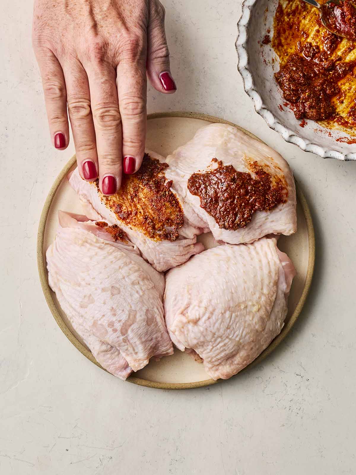 A plate of chicken thighs, with a hand rubbing an oily spice mix over the skins.