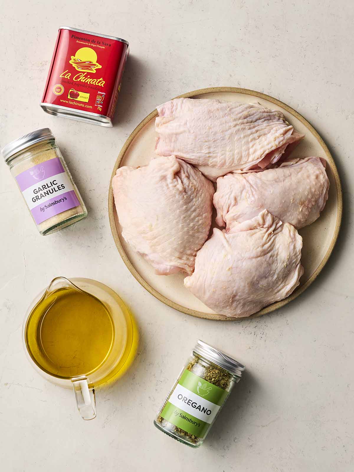 Ingredients laid out on a white counter, including a plate of four chicken thighs with skin on, a jug of oil, and paprika, garlic granules and oregano.