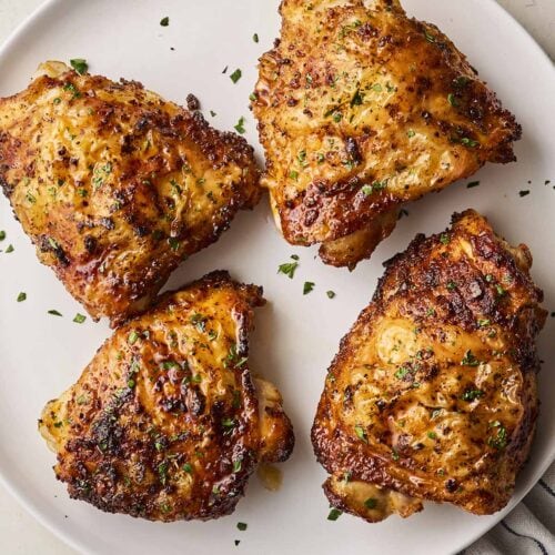 A white plate with four cooked chicken thighs on, coated in a marinade.