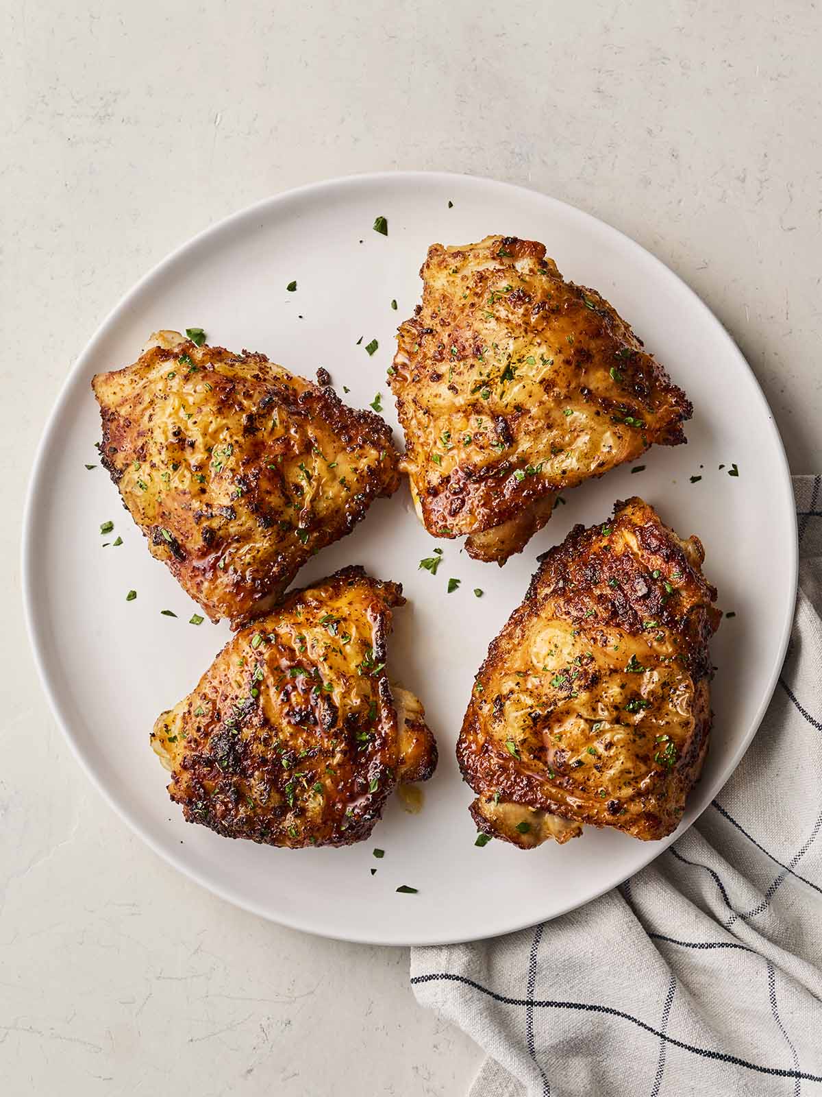 A white plate on a white surface with a tea towel and 4 chicken thighs ready to eat.