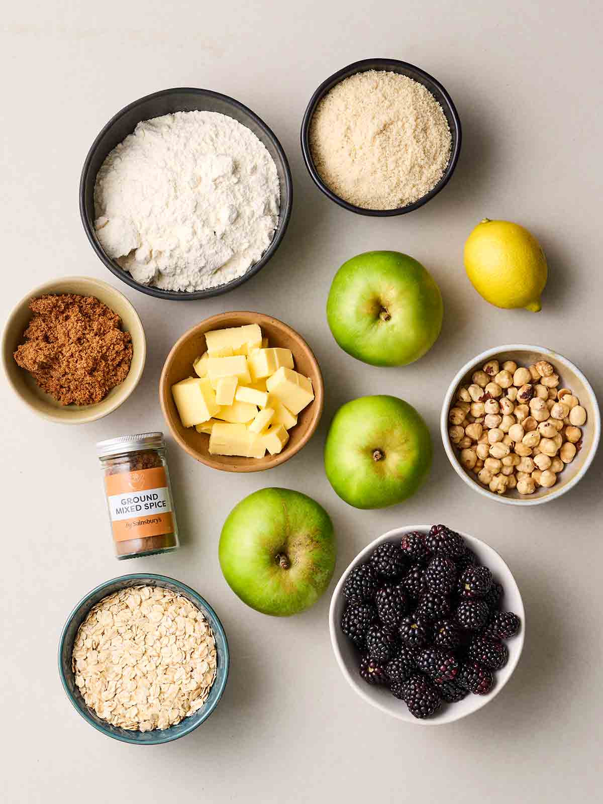 Ingredients laid out on a white counter, including apples, blackberries, flour, lemon, oats, butter, ground mixed spice, brown sugar and nuts.