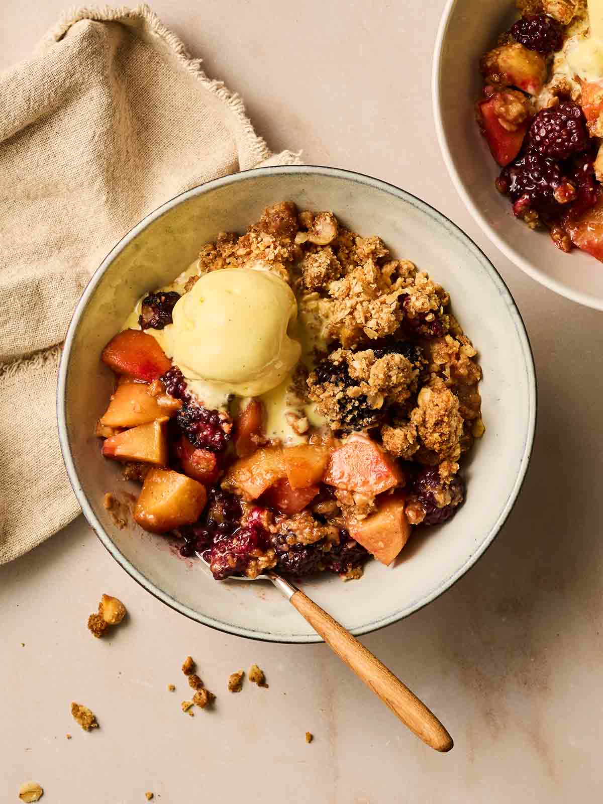A bowl filled with Apple and Blackberry Crumble with a scoop of vanilla ice cream and a spoon to eat with.