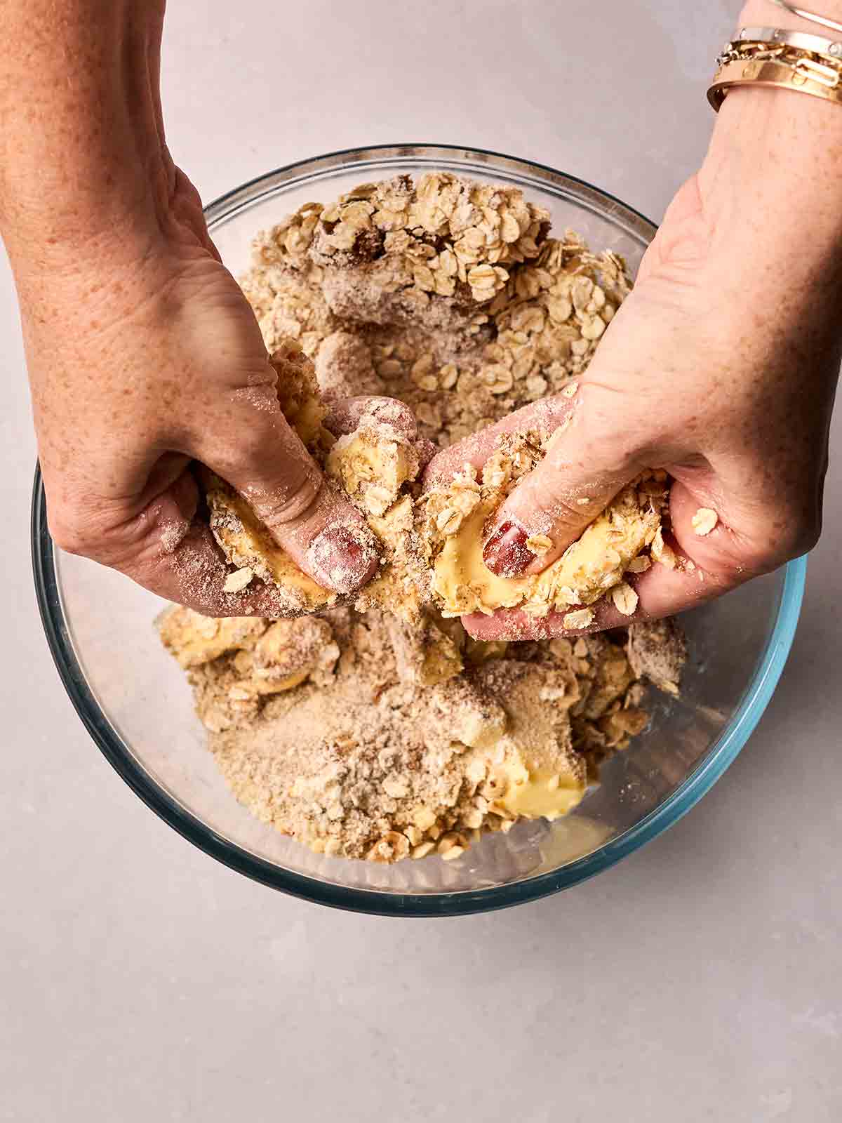 Two hands making a crumble topping over a glass bowl.