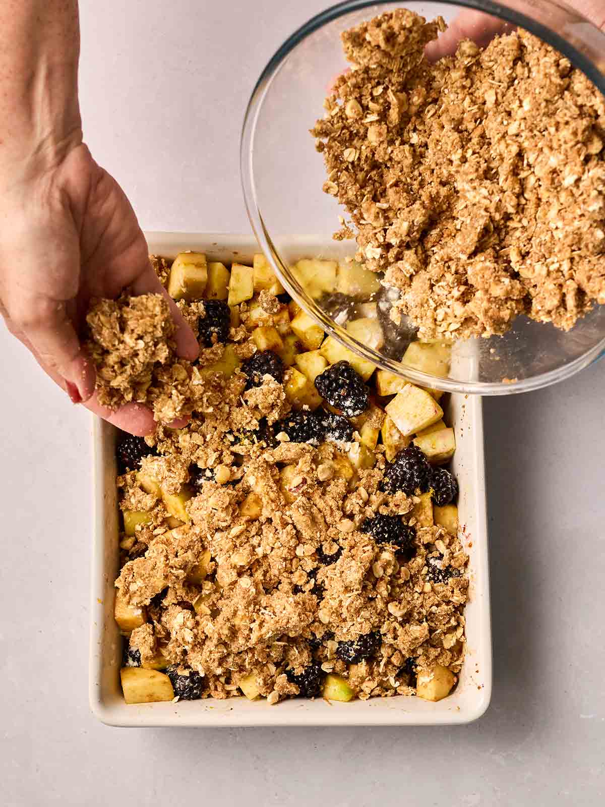 Crumble topping being scattered over apples and blackberries in a dish with hands from a glass bowl.