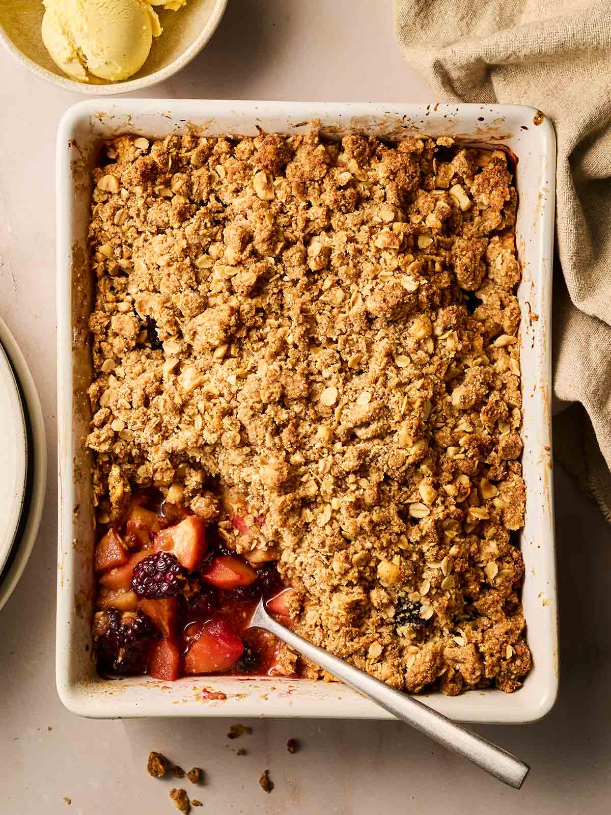A cooked Apple and Blackberry Crumble in a dish, being served up with a spoon on a table.