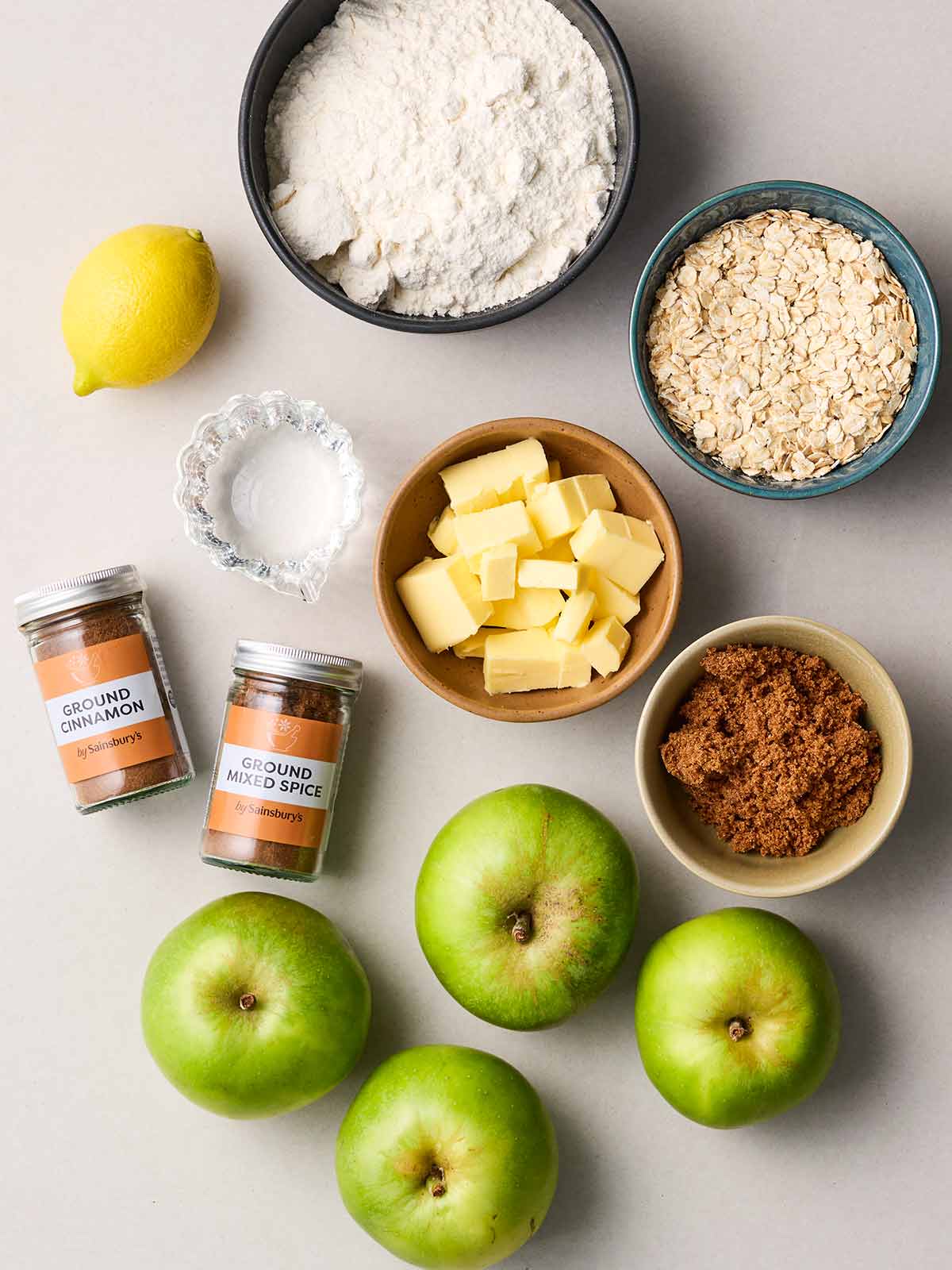 The ingredients for an Apple Crumble laid out in bowls on a white counter.