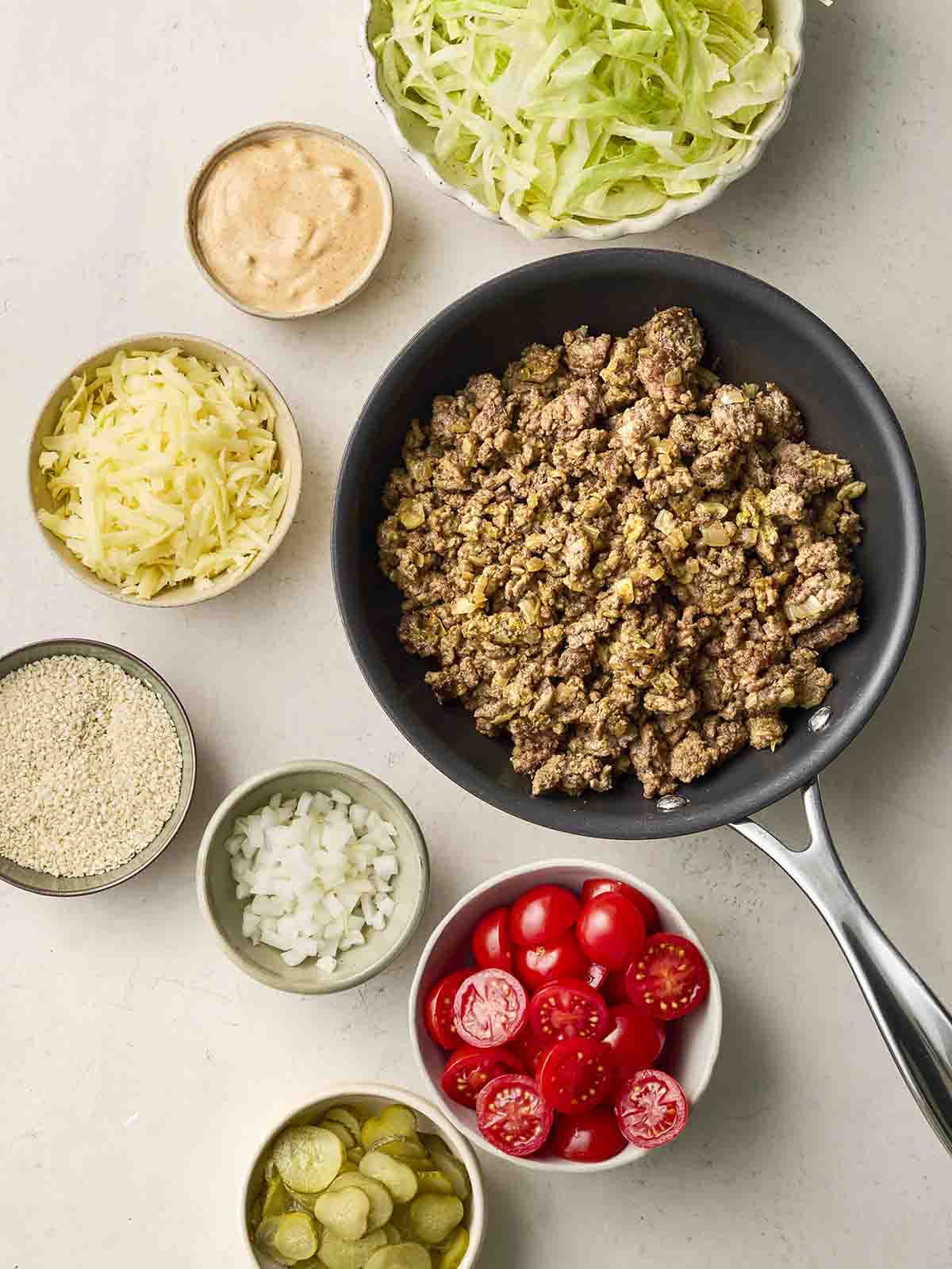 The ingredients for making Big Mac Bowls on a white counter, including ground beef, lettuce, sauce, grated cheese, onion, tomatoes and pickles.