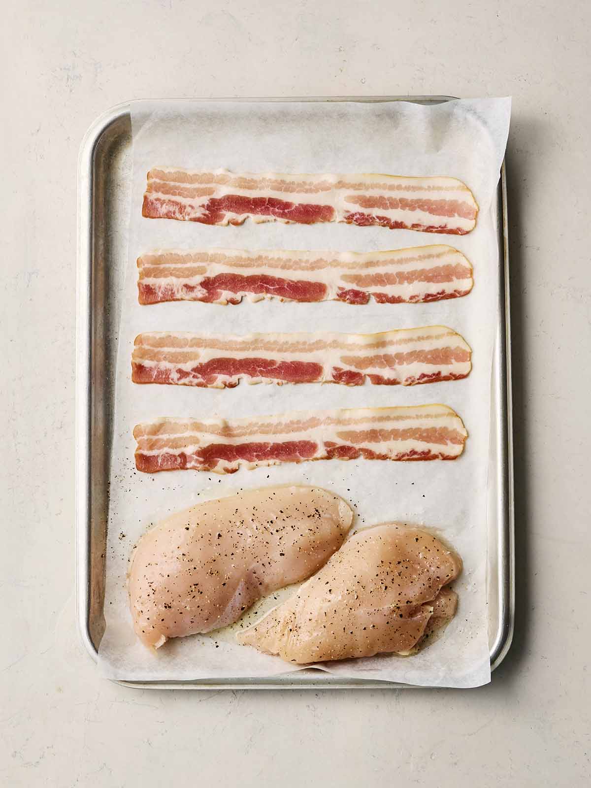 A baking tray with two raw chicken breasts and uncooked rashers of bacon on.