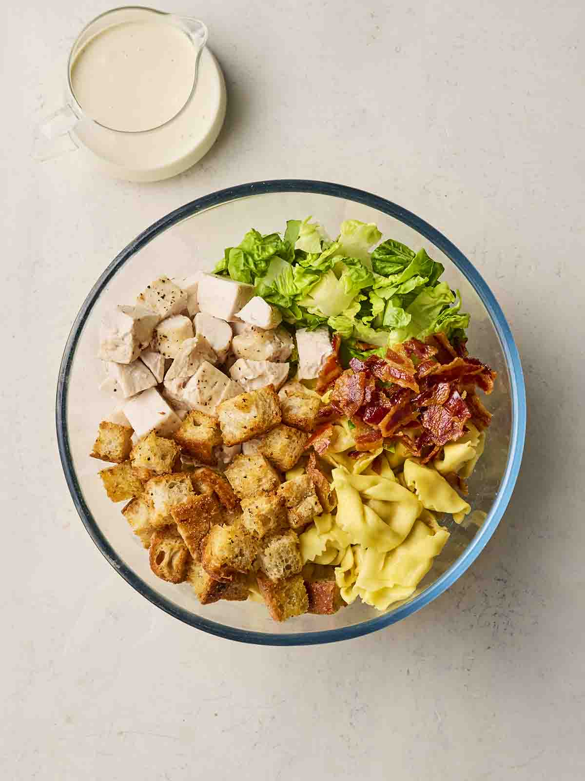 Ingredients in a bowl, including chicken, croutons, lettuce, pasta, bacon and a jug of caesar dressing at the side.