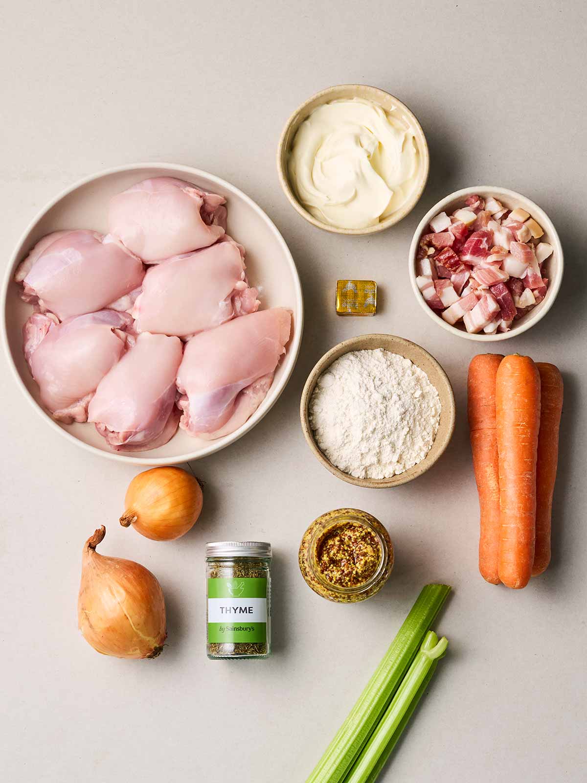 Raw ingredients laid out on a table, including chicken thighs, flour, carrots, bacon, onions, celery, time, a stock cube, mustard and yoghurt.