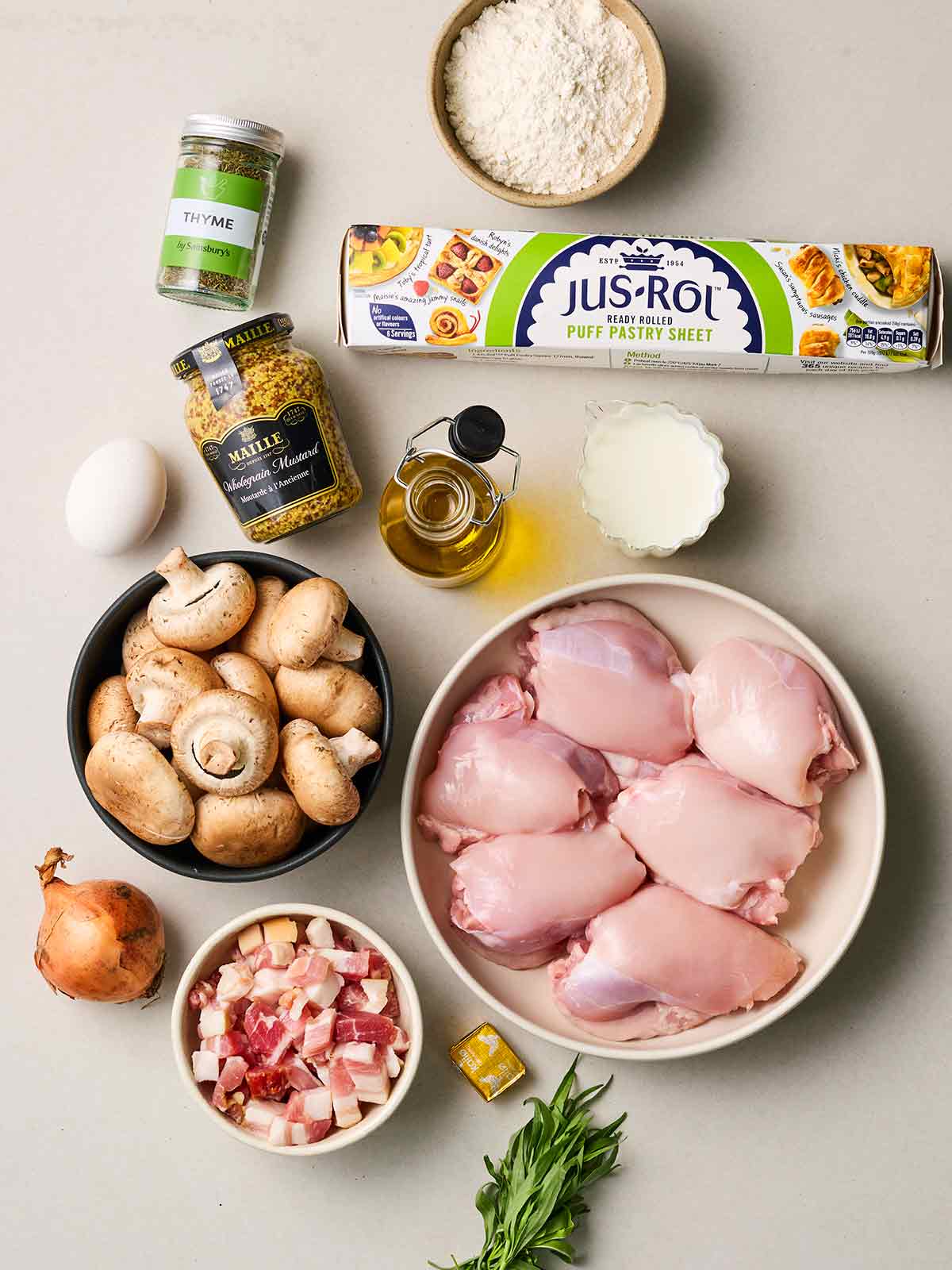 Raw ingredients laid out on a white counter, including chicken thighs, mushrooms, pastry, bacon, mustard, onion, thyme, oil and an egg.