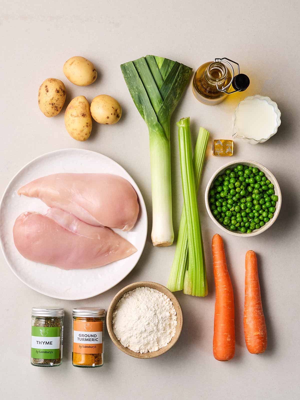Raw ingredients laid out on a white counter, including chicken, a leek, peas, carrot, potatoes and herbs.