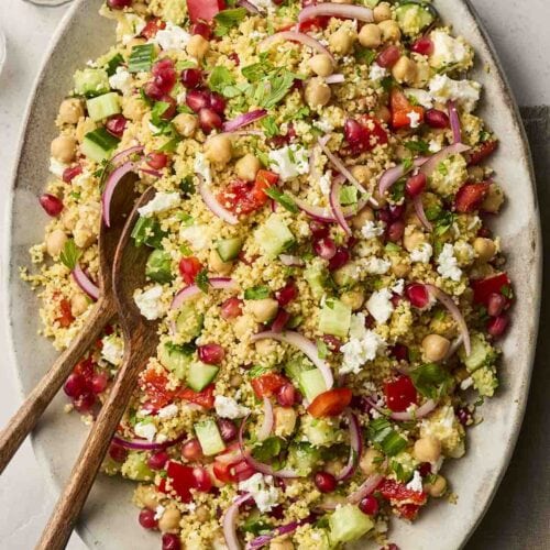 A colourful plate of salad with couscous, chickpeas and lots of other ingredients, with a pepper mill to the side and salad utensils to serve.