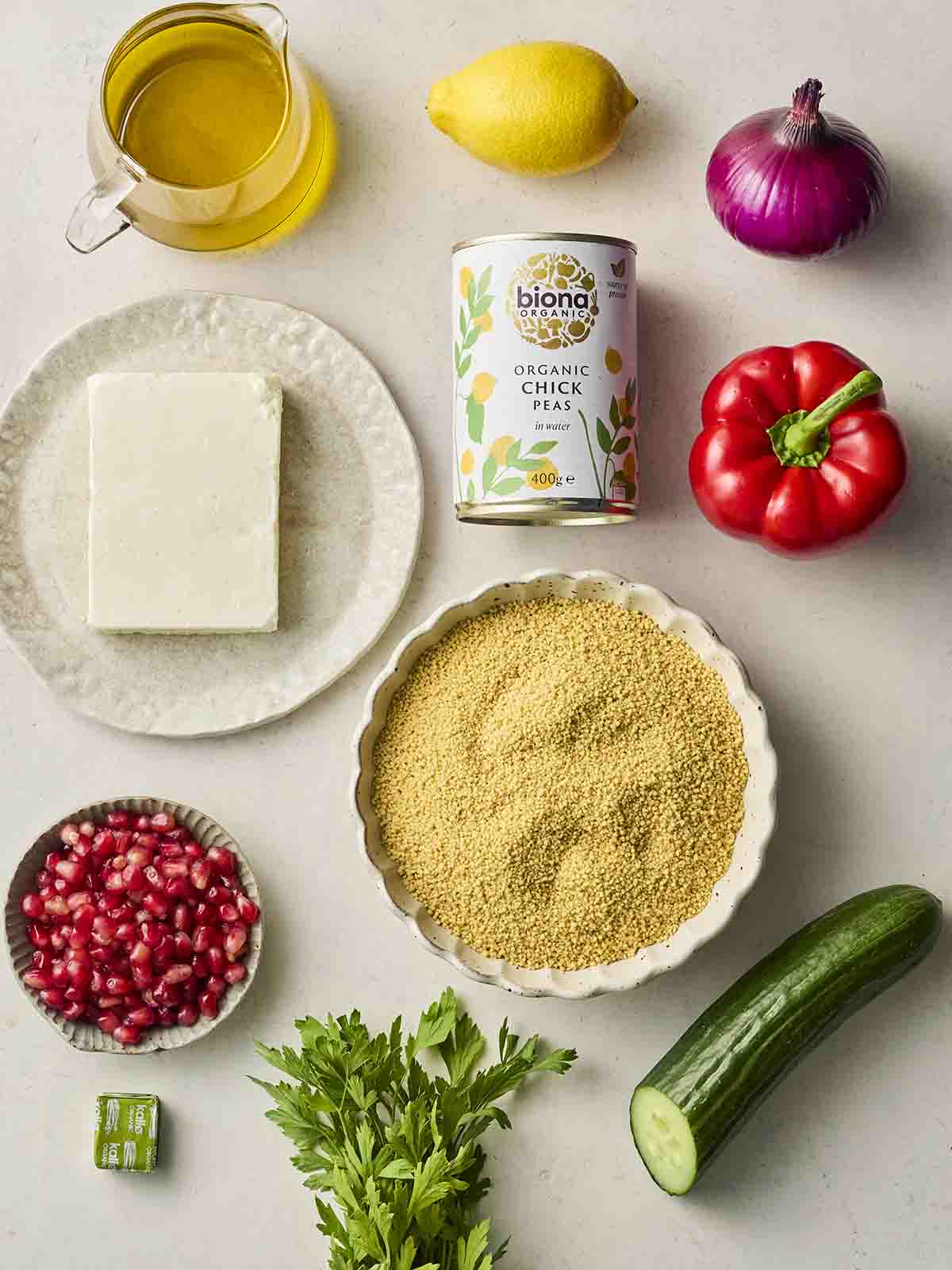 Ingredients laid out on a white counter, including chickpeas, feta, couscous, cucumber, red pepper, red onion, pomegranate seeds, lemon, oil and parsley.