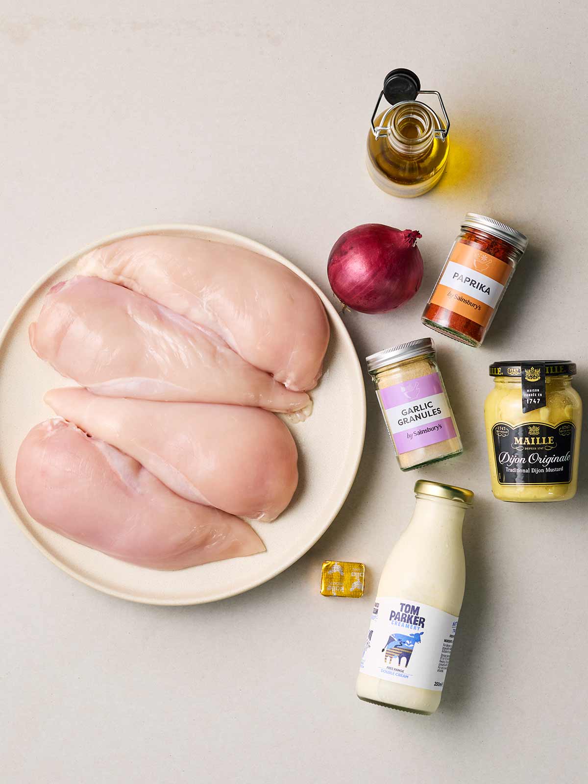 Raw ingredients laid out on a table, including chicken breasts, oil, paprika, mustard, cream and onion.